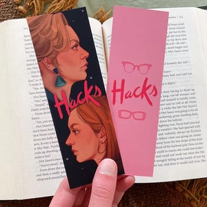 Könnte beinhalten: Zwei Lesezeichen mit dem Wort "Hacks". Eines ist schwarz mit Illustrationen von zwei Frauen und dem Wort "Hacks" in Rot. Das andere ist rosa mit dem Wort "Hacks" und Brillenillustrationen.