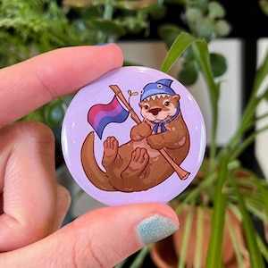 Pode incluir: Um botão redondo com uma lontra de desenho animado usando um chapéu azul e segurando uma bandeira do orgulho bissexual. A lontra é marrom com um fundo lilás claro. A bandeira é rosa, roxa e azul.