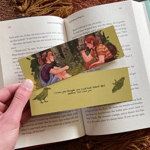 May include: Two bookmarks with illustrations of people in a forest setting. The top bookmark features a woman with long brown hair wearing a striped shirt and holding a camera. The bottom bookmark features a crow and the text "I know you thought you could hide behind that camera. But I saw you."