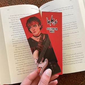 May include: Red bookmark with a drawing of a person with red hair and a guitar. The bookmark has the text "SEE YOU IN HELL" and a lightning bolt design. The bookmark is placed in an open book.