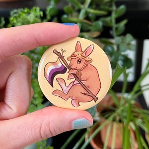 Op de afbeelding: Een ronde, lichtgele knoop met een cartoonkonijn met een hoed en een vlag met paarse, witte en gele strepen. Het konijn is bruin met een lichtbruine hoed en zit. De knoop wordt door een hand vastgehouden.