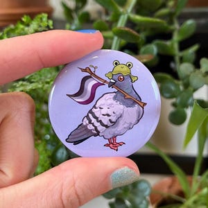 Pode incluir: Um botão redondo e lavanda com um pombo de desenho animado usando um chapéu de sapo e carregando um pau. O pombo tem uma bandeira ace presa ao pau. O botão é segurado pelos dedos de uma pessoa, com plantas verdes ao fundo.