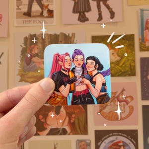 May include: A square sticker featuring three women with pink, purple, and black hair, forming a heart shape with their hands. The women are wearing stylish outfits with black and yellow accents. The sticker is held by a hand against a background of other stickers.