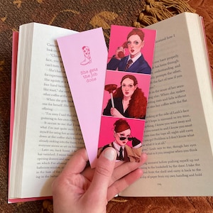 Pode incluir: Um conjunto de três marcadores de página cor-de-rosa com ilustrações de uma mulher em diferentes cenas. O texto no marcador de página diz "She gets the job done".