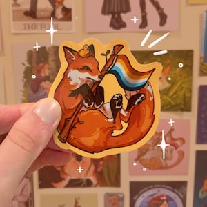 May include: A sticker featuring an orange fox with a crown, holding a flag with rainbow colors. The fox is lying down, with a whimsical design. The sticker has a yellow border and is held by a hand.