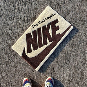 May include: Rectangular beige rug with a brown Nike logo and the text "The Rug Legare". The rug is placed on a gray concrete surface. The Nike swoosh and lettering are in a dark brown color.