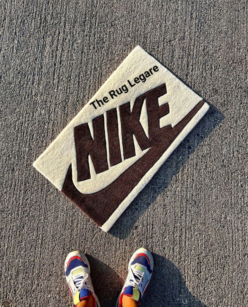 May include: Rectangular beige rug with a brown Nike logo and the text "The Rug Legare". The rug is placed on a gray concrete surface. The Nike swoosh and lettering are in a dark brown color.