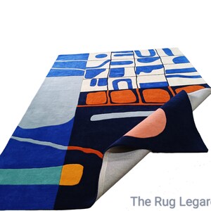 May include: A rectangular rug with an abstract design. The rug features a colour palette of blues, orange, and cream. The design includes geometric shapes and curved lines. The rug is made of a soft material and has a folded corner.