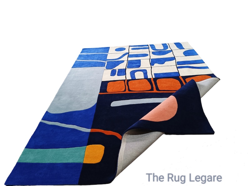 May include: A rectangular rug with an abstract design. The rug features a colour palette of blues, orange, and cream. The design includes geometric shapes and curved lines. The rug is made of a soft material and has a folded corner.
