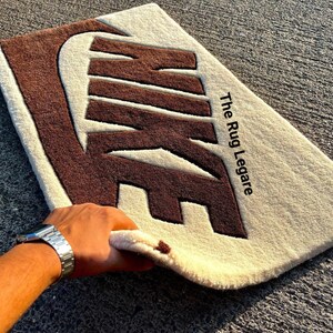 May include: A rectangular rug featuring the Nike logo in brown against a cream background. The text "The Rug Legare" is printed in black. The rug is made of a soft, textured material.