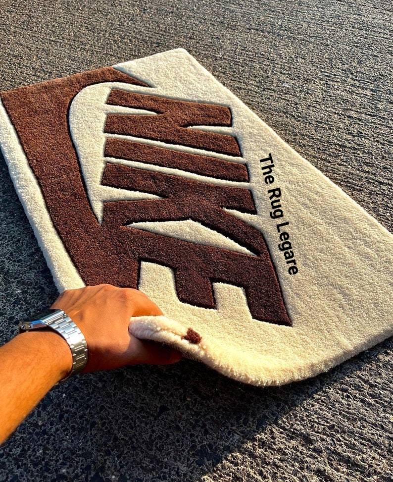 May include: A rectangular rug featuring the Nike logo in brown against a cream background. The text "The Rug Legare" is printed in black. The rug is made of a soft, textured material.