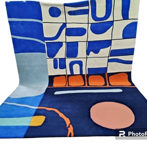 May include: An abstract rug with a modern design in blue, orange, and cream. The rug has a dark blue base with a lighter blue and cream-coloured pattern. An orange border and a peach-coloured circle add to the design.