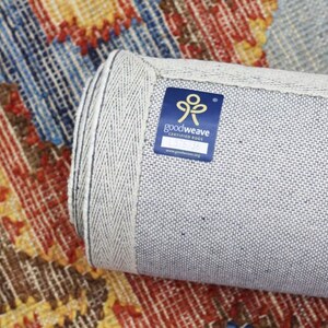 May include: Rolled-up area rug with a colourful geometric pattern in shades of red, blue, yellow, and brown. The rug's edge is visible, showing a light grey backing and a blue GoodWeave certified rug label.