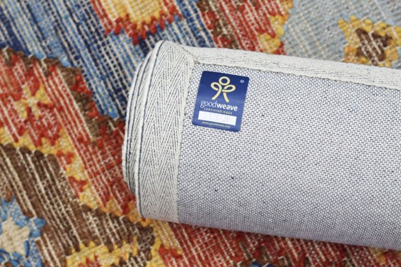 May include: Rolled-up area rug with a colourful geometric pattern in shades of red, blue, yellow, and brown. The rug's edge is visible, showing a light grey backing and a blue GoodWeave certified rug label.