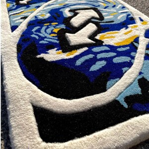 May include: A close-up of a rug with a design featuring a blue and white starry night sky with a large white arrow. The rug has a white border and a black area. The text "The Rug Legare" is at the bottom.