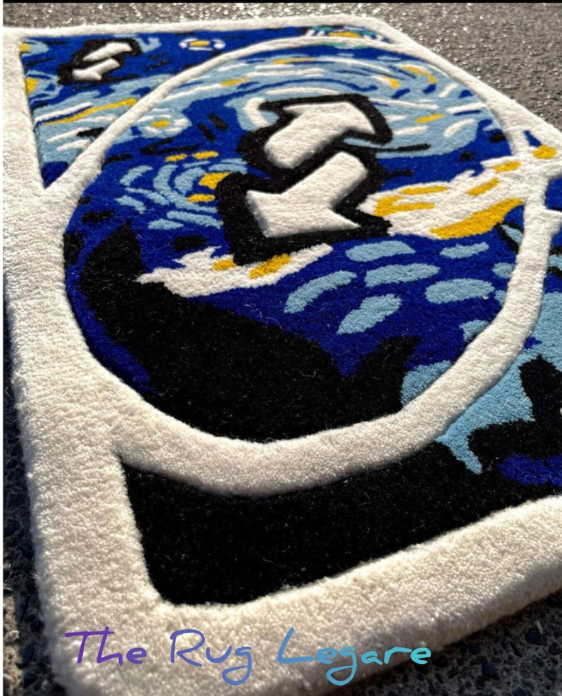 May include: A close-up of a rug with a design featuring a blue and white starry night sky with a large white arrow. The rug has a white border and a black area. The text "The Rug Legare" is at the bottom.