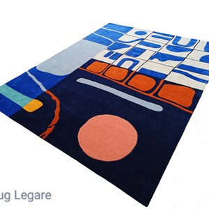 May include: A large, rectangular rug with an abstract design. The rug features a navy blue base with sections of light blue, orange, and white. A large peach-coloured circle and a light grey line are also present. The text "The Rug Legare" is at the bottom.