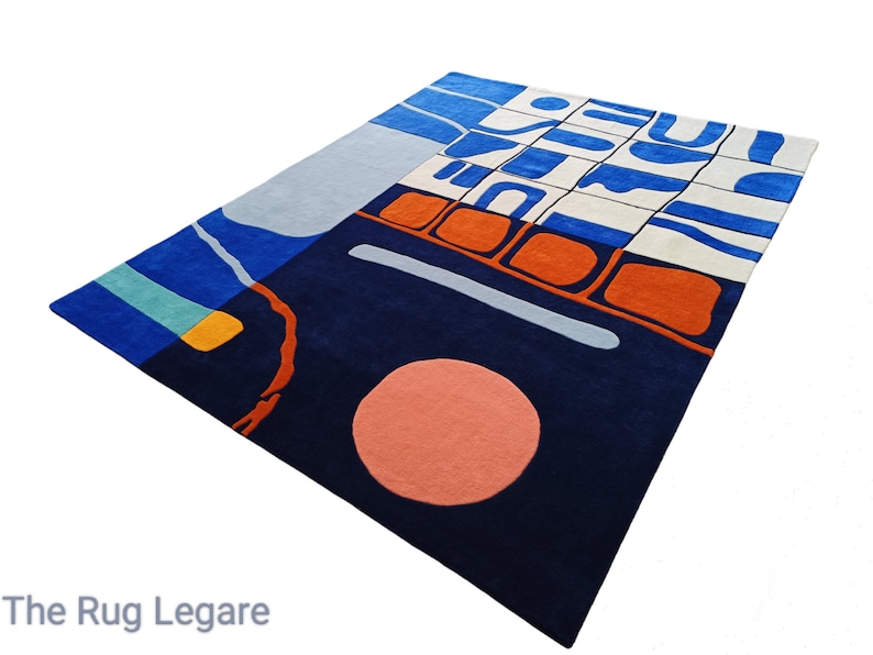 May include: A large, rectangular rug with an abstract design. The rug features a navy blue base with sections of light blue, orange, and white. A large peach-coloured circle and a light grey line are also present. The text "The Rug Legare" is at the bottom.