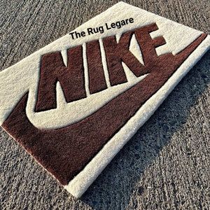 May include: Rectangular rug with the Nike logo in brown against a cream background. The text "The Rug Legare" is in the upper left corner. The rug is placed on a textured gray surface.