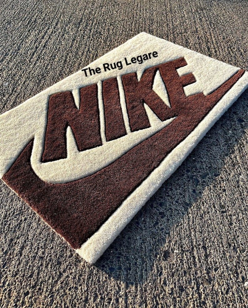May include: Rectangular rug with the Nike logo in brown against a cream background. The text "The Rug Legare" is in the upper left corner. The rug is placed on a textured gray surface.