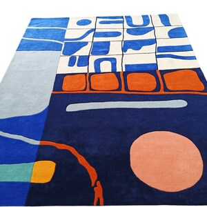 May include: Modern geometric rug in navy blue, light blue, orange, and white. The rug has an abstract design with a large peach circle and other shapes. The rug is rectangular in shape and would suit a living room.