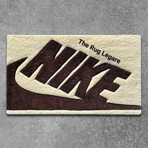 May include: A rectangular rug with the Nike logo in dark brown against a cream-colored background. The text "The Rug Legare" is printed in black. The rug is placed on a gray surface.
