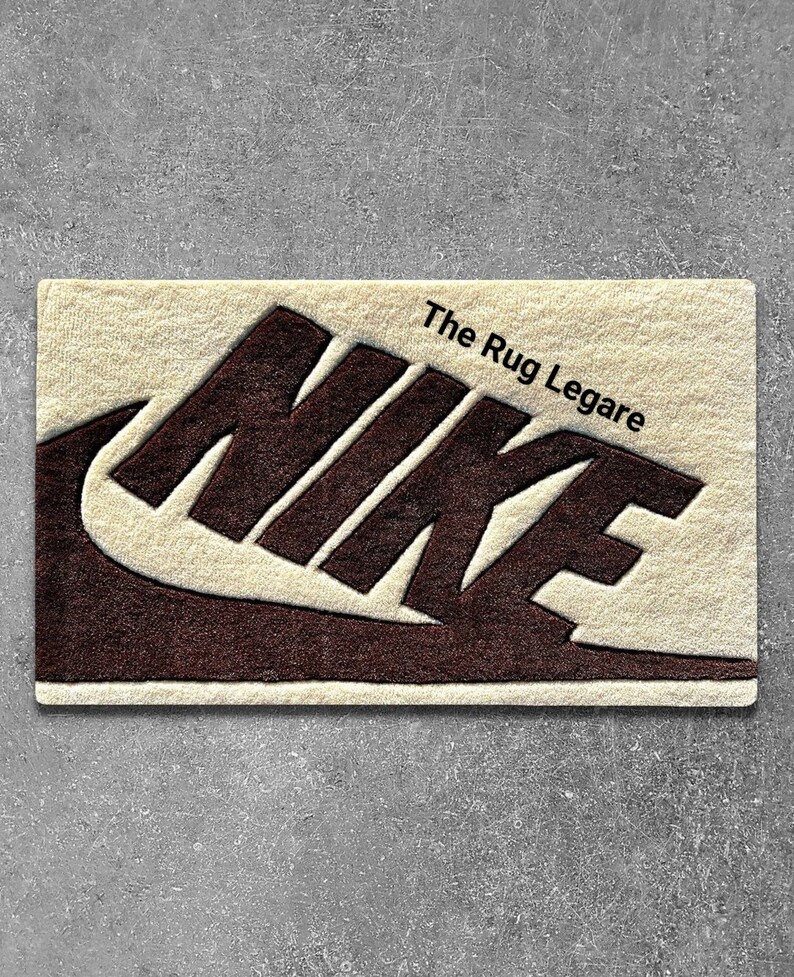 May include: A rectangular rug with the Nike logo in dark brown against a cream-colored background. The text "The Rug Legare" is printed in black. The rug is placed on a gray surface.