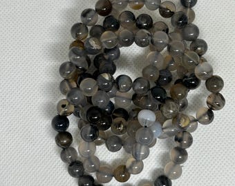 May include: Several strands of beaded bracelets. The beads are round and vary in shades of gray, black, and white. The bracelets are arranged in a loose, overlapping pattern against a white background.