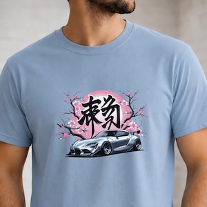 Könnte beinhalten: Ein hellblaues Kurzarm-T-Shirt mit einem Grafikdruck eines silbernen Sportwagens vor einem rosa Kreis und Kirschblütenzweigen, mit großen schwarzen japanischen Kanji-Schriftzeichen über dem Fahrzeug.