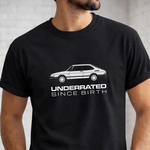 May include: A black short-sleeved t-shirt featuring a white line art illustration of a classic hatchback car centered on the chest. Below the car graphic, the text reads "UNDERRATED SINCE BIRTH" in a clean, bold, white sans-serif typeface.