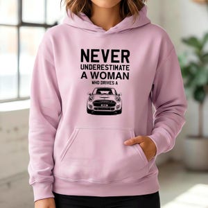 May include: A pale pink hoodie with a front pocket and a drawstring hood. The front of the hoodie features the text "NEVER UNDERESTIMATE A WOMAN WHO DRIVES A" above a black and white illustration of a Mini Cooper.