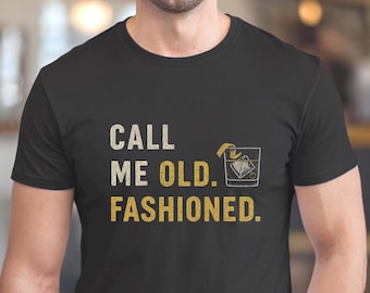 May include: A black short-sleeved t-shirt featuring the text "CALL ME OLD. FASHIONED." in a distressed font. The word "OLD." and "FASHIONED." are in gold, while "CALL ME" is in white. A graphic of an Old Fashioned cocktail is included.