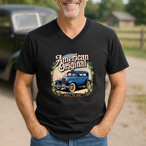 May include: Black V-neck t-shirt featuring a vintage blue car graphic with the words "American Original" and "Built to Last." The design is surrounded by a decorative border. The shirt is made of a soft material.