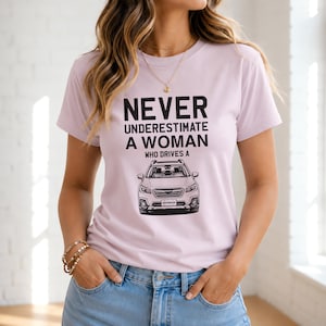 May include: A light purple short-sleeved t-shirt featuring black text that reads NEVER UNDERESTIMATE A WOMAN WHO DRIVES A above a black and white graphic illustration of a Subaru Outback vehicle with the word OUTBACK on the license plate.