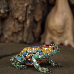 Puede incluir: Escultura de rana de mosaico colorida con un patrón detallado de varias formas y colores, incluyendo verde, azul, amarillo y rojo. La rana está posada sobre una superficie marrón, mostrando su intrincado diseño y artesanía artística.