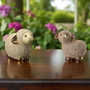 May include: Two ceramic sheep figurines. The sheep have a beige body with a textured wool appearance, cream-colored faces, and gold accents on the ears and hooves. They are posed on a dark wooden surface with a blurred floral background.