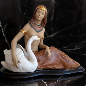 Może przedstawiać: Ceramiczna figurka kobiety z rudymi włosami, w złotej opasce i bransoletach oraz ozdobnym kołnierzu. Siedzi obok białego łabędzia. Kobieta ma na sobie brązową spódnicę. Figurka stoi na czarnej podstawie.