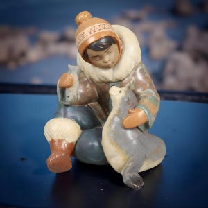 Può includere: Una statuina in ceramica raffigurante una persona in abiti invernali, seduta e che abbraccia una foca. La figura indossa un cappello marrone, un parka color crema e pantaloni blu. La foca è grigia e color crema. La statuina ha un'espressione serena e tiene un pesce.