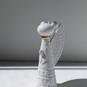May include: A white ceramic angel figurine with a stylized design. The angel has a round head with two large ears, a gold-coloured collar, and detailed wings. The base features a heart pattern.