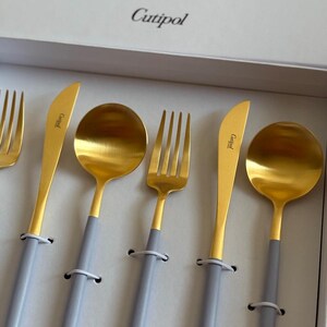 May include: A set of Cutipol flatware in a white box. The utensils have gold-colored heads and light gray handles. The set includes forks, knives, and spoons. The Cutipol logo is visible on the box and the knife.