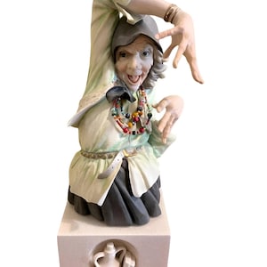 May include: A ceramic figurine of a woman with a mischievous expression, wearing a hat, a light green blouse, and a dark skirt. The figurine has a colourful beaded necklace and is mounted on a square base with a carved design.