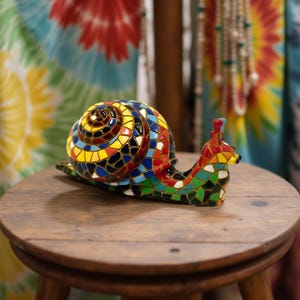 Op de afbeelding: Een kleurrijk mozaïek slakfiguur met een spiraalvormig huis in geel, blauw, rood en groen. De slak staat op een kleine, ronde houten kruk. De achtergrond toont een tie-dye stof.