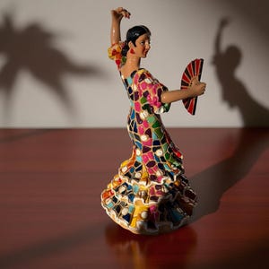 May include: A ceramic flamenco dancer figurine in a mosaic-patterned dress, holding a red and black fan. The dancer's pose is dynamic, with one arm raised. The figurine is on a wooden surface.