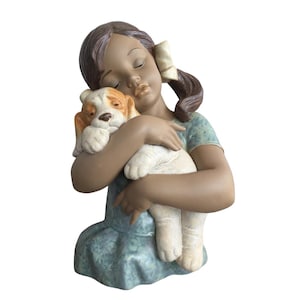 May include: A ceramic figurine depicting a girl in a blue dress cuddling a puppy. The girl has brown hair and a white bow. The puppy is white and tan. A decorative ornament for the home.
