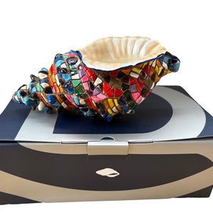 Puede incluir: Una escultura de concha marina de mosaico colorido con un interior color crema, apoyada sobre una caja con estampado azul marino y blanco. La concha está adornada con una vibrante variedad de pequeñas baldosas multicolores, creando una pieza decorativa única.