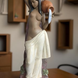Può includere: Statuina in ceramica di una donna che versa acqua da un boccale di terracotta. La figura è drappeggiata in un panno bianco e poggia su una base con accenti floreali. La donna ha i capelli scuri e la carnagione chiara.