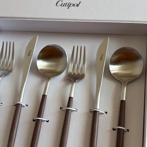 May include: A set of Cutipol silverware in a white box. The set includes forks, knives, and spoons with silver heads and dark brown wooden handles. The Cutipol logo is visible on the box and the knife blades.