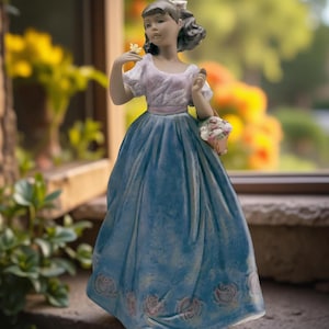 Può includere: Statuina di porcellana di una giovane donna in un lungo abito blu con rose rosa. Tiene in mano un fiore e un cestino di fiori. La statuina ha un fiocco bianco tra i capelli e un top rosa e bianco. Oggetto decorativo.