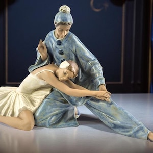 Può includere: Statuina in porcellana raffigurante due ballerini di danza classica. Una ballerina è inginocchiata con un tutù bianco, mentre l'altra, in costume blu, sembra sostenerla. La scultura ha una finitura opaca ed è ambientata su uno sfondo blu scuro.