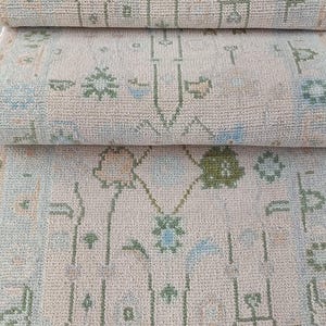 May include: A folded area rug featuring a cream-colored base with a repeating pattern of small floral and geometric motifs in muted shades of sage green, light blue, and soft peach. The rug has a textured pile and a fringed edge at the top.
